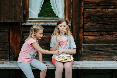 Auf diesem Bild sitzen zwei Mädels auf einer Holzbank mit einem Teller voller Kaiserschmarren in der Hand, sie teilen ihn sich | © Tirol Werbung | Verena Kathrein